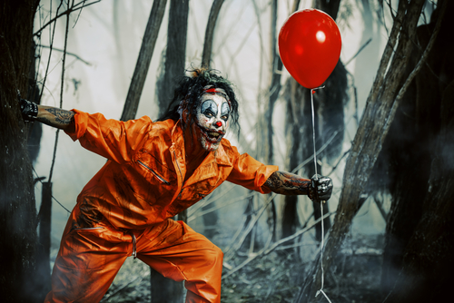 Scary clown holding red balloon in forest