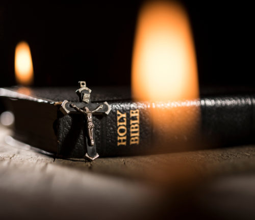 Cross leaning against a bible with candle flames against a dark background