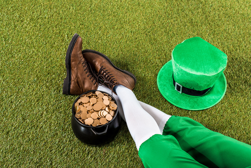 Cropped view of a Leprechaun relaxing on grass with his legs near his pot of gold on one side and hat on the other.