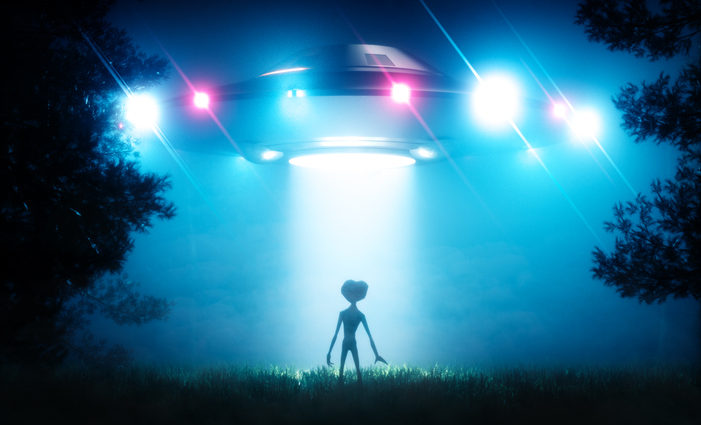 Silhouette of alien standing in beam of light from UFO hovering above the ground.