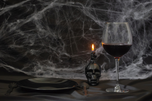 Scary halloween dinner party concept with a glass of red wine, black plate, knife, fork and skull candle with cobweb on black background.